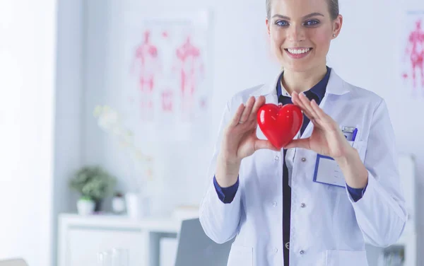 Mujer joven doctora sosteniendo un corazón rojo, de pie sobre el fondo del hospital — Foto de Stock