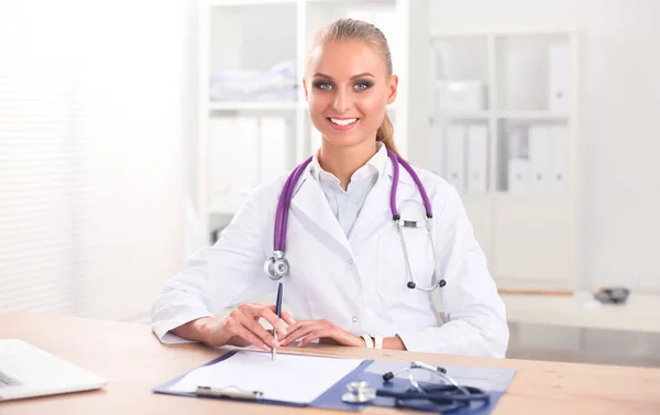 Mooie jonge glimlachende vrouwelijke arts zitten aan het bureau en schrijven. — Stockfoto