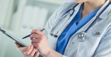 Steteskoplu bir doktor elinde bir defter tutuyor. Hastanede ayakta dikilirken panoda tıbbi form dolduran bir kadın doktorun yakın çekimi.