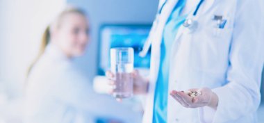 Doktorun arka planda meslektaşıyla klinikte elinde haplar ve bir bardak su tutarken görüntüsü..