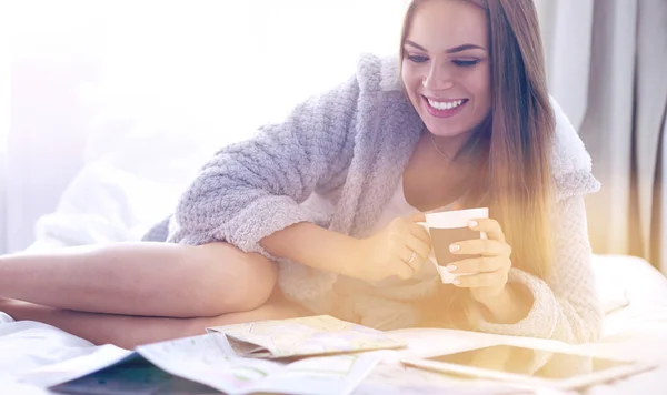 Avslappnad ung kvinna sitter på sängen med en kopp kaffe och digital tablett — Stockfoto