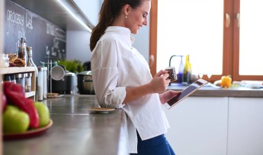 Genç bir kadın mutfakta tablet kullanıyor ve kahve içiyor.