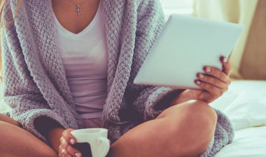 Bir fincan kahve ve dijital tabletle yatakta oturan rahat genç bir kadın.