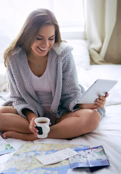 Mujer joven y relajada sentada en la cama con una taza de café y una tableta digital —  Fotos de Stock