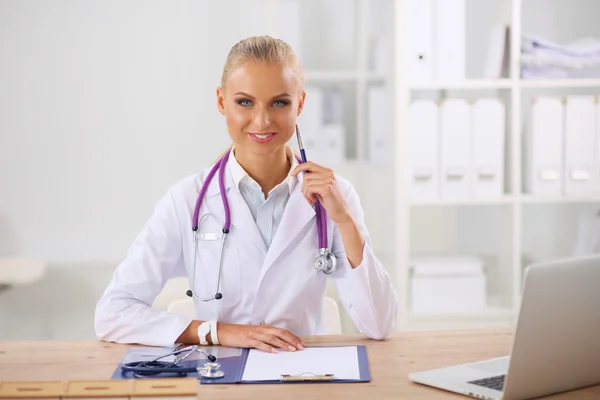 Schöne junge lächelnde Ärztin sitzt am Schreibtisch und w — Stockfoto
