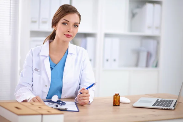 Schöne junge lächelnde Ärztin sitzt am Schreibtisch und w — Stockfoto