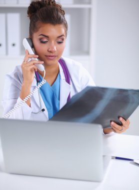 Röntgen taraması yapan kadın doktor teşhis merkezinde telefonla konuşuyor, masada oturuyor.