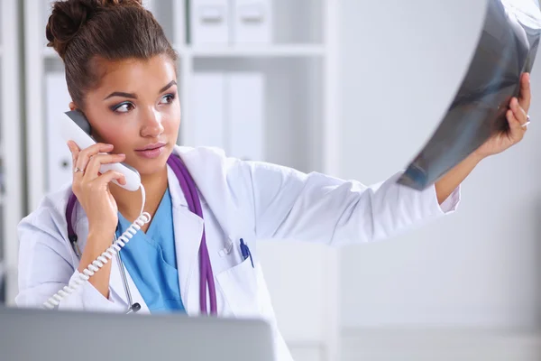 Médico femenino que mira la radiografía y habla por teléfono en el centro de diagnóstico, sentado en el escritorio —  Fotos de Stock