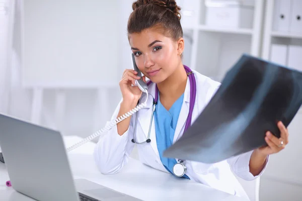 Médico femenino que mira la radiografía y habla por teléfono en el centro de diagnóstico, sentado en el escritorio —  Fotos de Stock