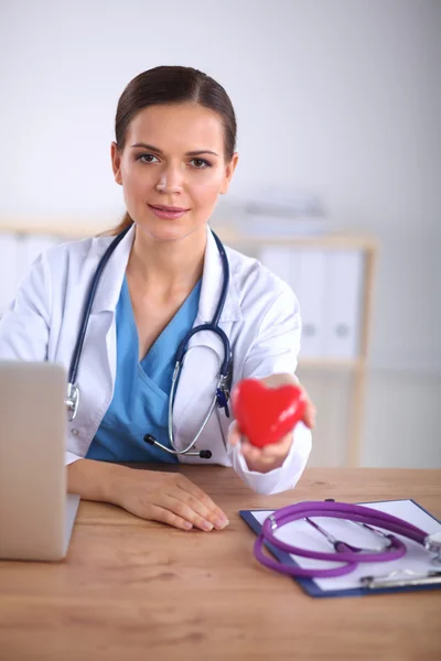 Young doctor with red heart symbol sitting at desk isolated — Stock Photo, Image