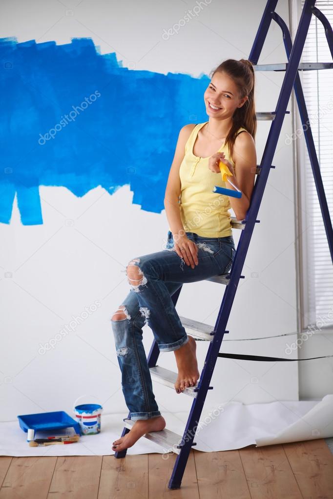 Happy beautiful young woman doing wall painting, sitting on ladder