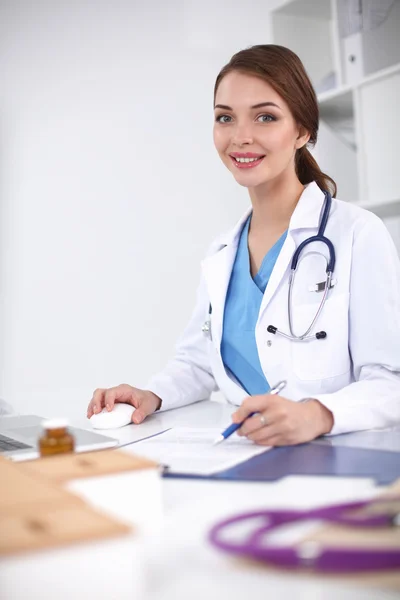 Hermosa joven sonriente doctora sentada en el escritorio y escribiendo. — Foto de Stock