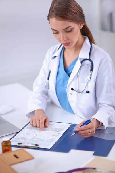 Hermosa joven sonriente doctora sentada en el escritorio y escribiendo. — Foto de Stock