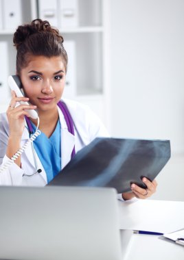 Röntgen taraması yapan kadın doktor teşhis merkezinde telefonla konuşuyor, masada oturuyor.