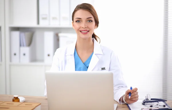 Hermosa joven sonriente doctora sentada en el escritorio y escribiendo. — Foto de Stock