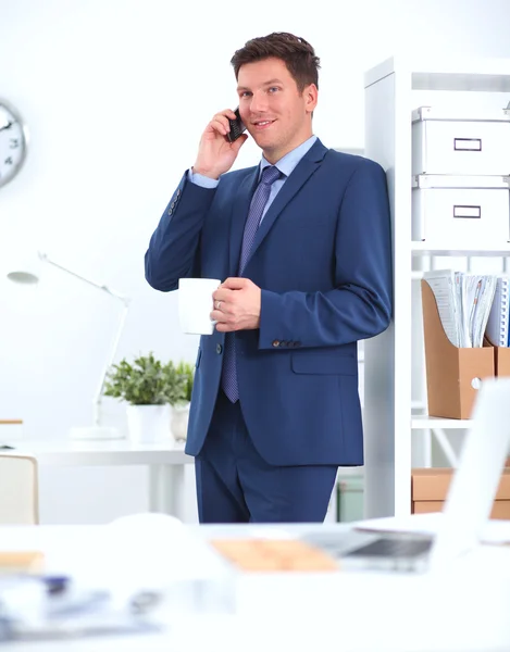 Hombre de negocios sonriente de pie y usando el teléfono móvil en la oficina — Foto de Stock