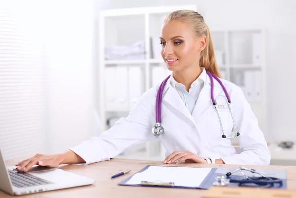 Hermosa joven sonriente doctora sentada en el escritorio y escribiendo. — Foto de Stock