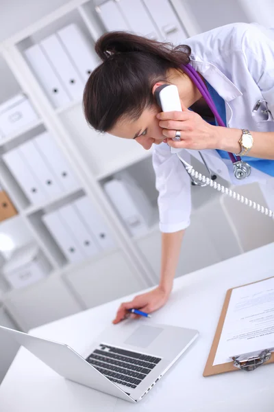 Young woman doctor in white coat at computer using phone — Stock Photo, Image