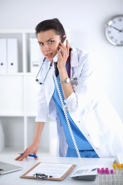 Young woman doctor in white coat at computer using phone — Stock Photo, Image