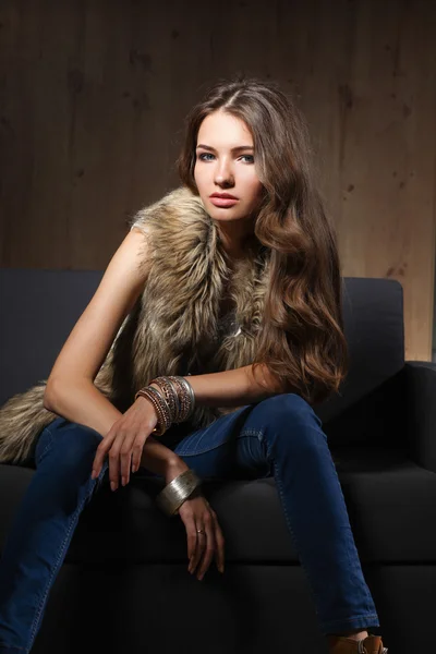 Retrato de una mujer elegante sentada en un sofá negro con pantalones vaqueros azules y chaleco de piel — Foto de Stock