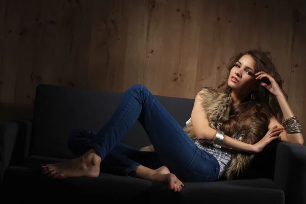 Retrato de una mujer elegante sentada en un sofá negro con pantalones vaqueros azules y chaleco de piel — Foto de Stock