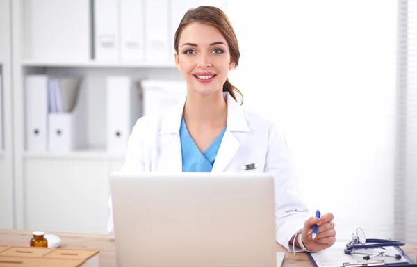 Beautiful young smiling female doctor sitting at the desk and writing. — Stock Photo, Image