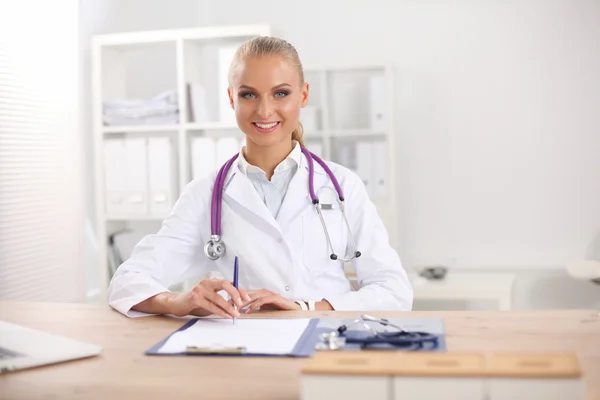Schöne junge lächelnde Ärztin sitzt am Schreibtisch und schreibt. — Stockfoto