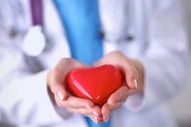 Kırmızı bir kalp taşıyan genç bir kadın doktor, gri arka planda duruyor.