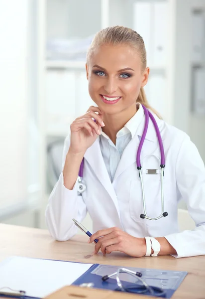 Schöne junge lächelnde Ärztin sitzt am Schreibtisch und schreibt. — Stockfoto