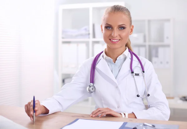 Mooie jonge glimlachende vrouwelijke arts zitten aan het bureau en schrijven. — Stockfoto