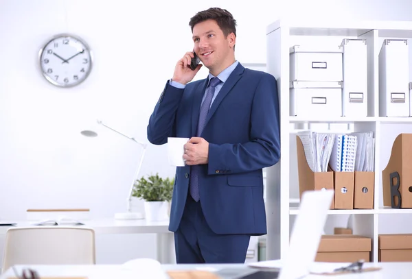 Hombre de negocios sonriente de pie y usando el teléfono móvil en la oficina —  Fotos de Stock