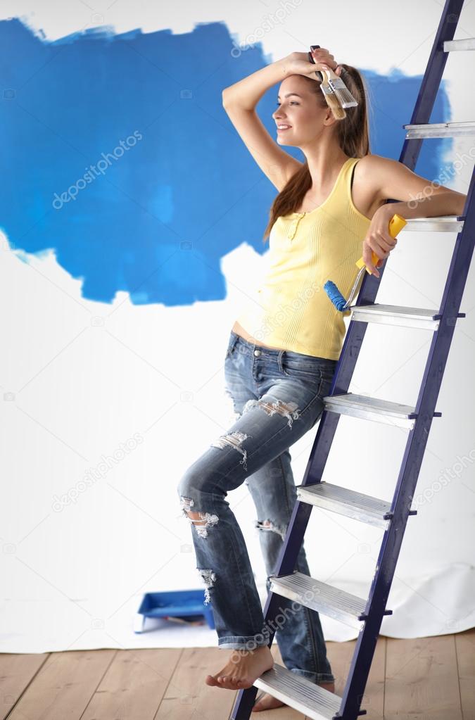 Happy beautiful young woman doing wall painting, standing near ladder