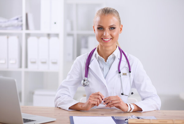 Beautiful young smiling female doctor sitting at the desk and writing.