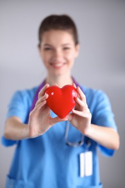 Kırmızı bir kalp taşıyan genç bir kadın doktor, gri arka planda duruyor.
