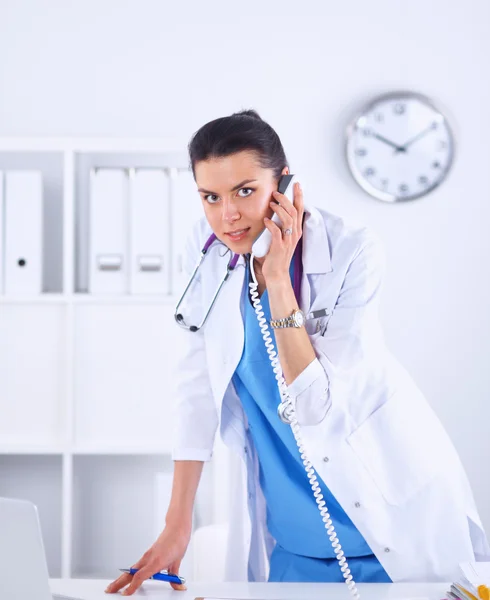 Young woman doctor in white coat at computer using phone — Stock Photo, Image
