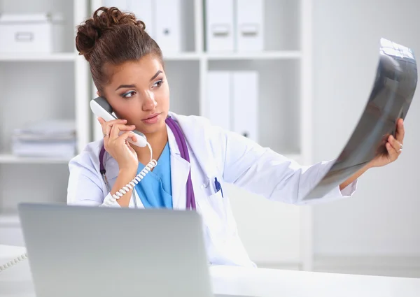 Médico femenino que mira la radiografía y habla por teléfono en el centro de diagnóstico, sentado en el escritorio — Foto de Stock