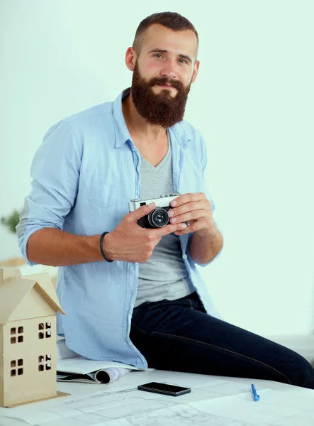 Portret van mannelijke ontwerper zittend op een tafel met blauwdrukken en vasthouden van camera — Stockfoto