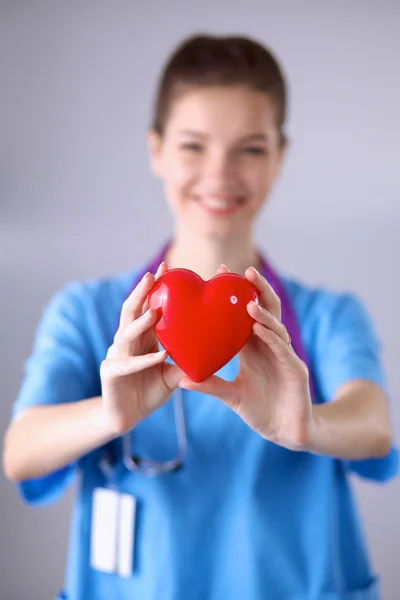 Νεαρή γυναίκα γιατρός κρατώντας μια κόκκινη καρδιά, στέκεται σε γκρι φόντο — Φωτογραφία Αρχείου