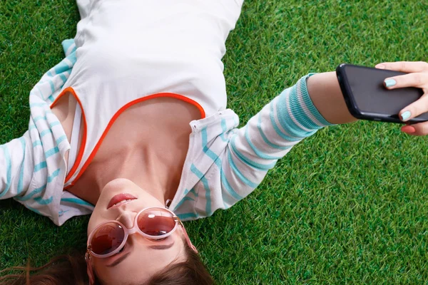 Mooie jonge vrouw selfie maken door haar telefoon liggend in groene gras — Stockfoto