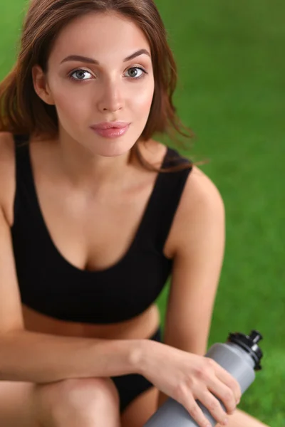 Young female athlete sitting on grass after exericise and holding a water bottle — Stock Photo, Image