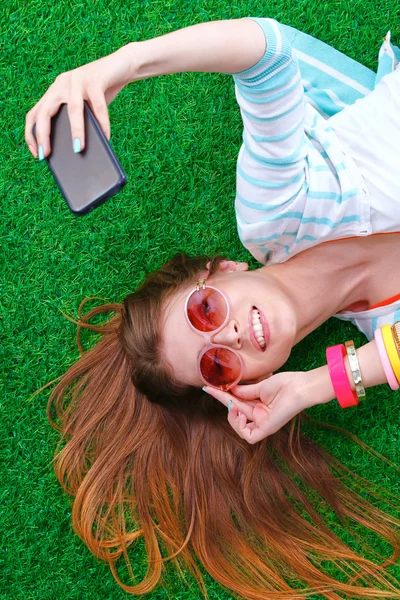 Bela jovem mulher fazendo selfie por seu telefone enquanto deitado na grama verde — Fotografia de Stock