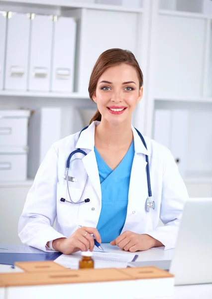 Hermosa joven sonriente doctora sentada en el escritorio y escribiendo. Fotos de stock libres de derechos