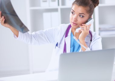 Röntgen taraması yapan kadın doktor teşhis merkezinde telefonla konuşuyor, masada oturuyor.