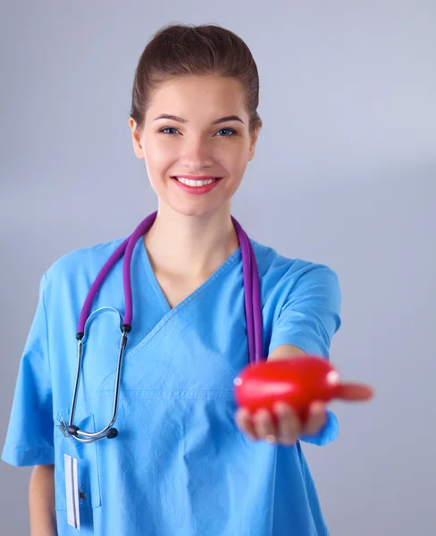 Mujer joven médico sosteniendo un corazón rojo, de pie sobre fondo gris — Foto de Stock