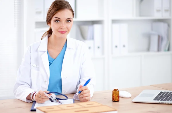 Hermosa joven sonriente doctora sentada en el escritorio y escribiendo. — Foto de Stock