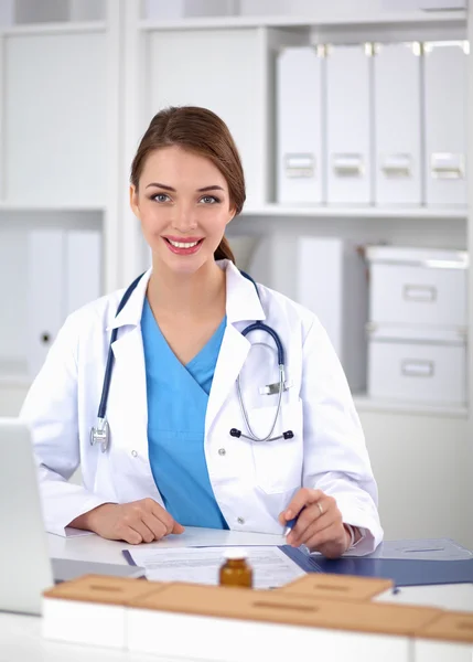 Hermosa joven sonriente doctora sentada en el escritorio y escribiendo. —  Fotos de Stock