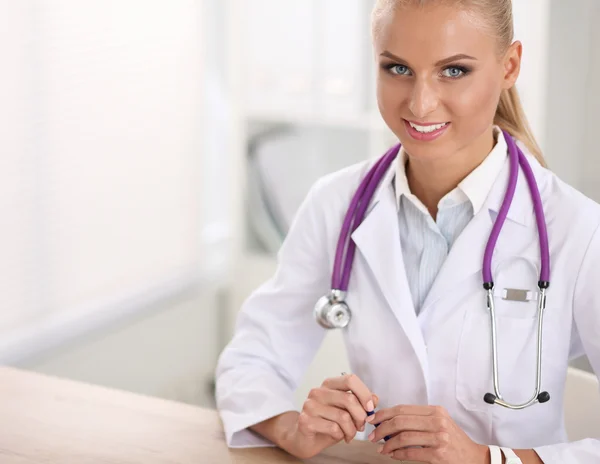 Bonito jovem sorridente médico feminino sentado na mesa e escrevendo. — Fotografia de Stock