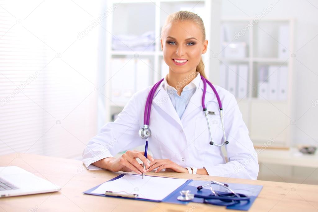 Beautiful young smiling female doctor sitting at the desk and writing.