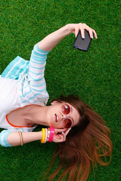 Mooie jonge vrouw selfie maken door haar telefoon liggend in groene gras — Stockfoto