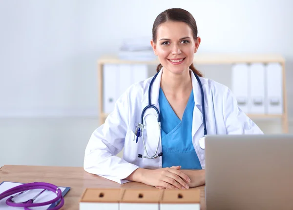 Bonito jovem sorridente médico feminino sentado na mesa e escrevendo. — Fotografia de Stock